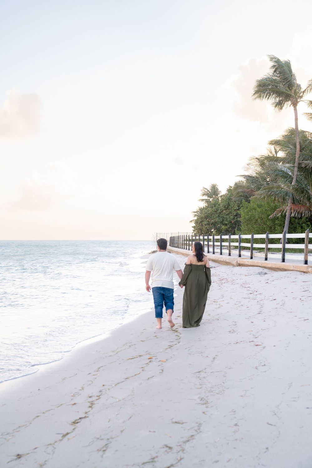 A couple walks hand in hand along a serene beach with palm trees and a wooden fence at sunset.