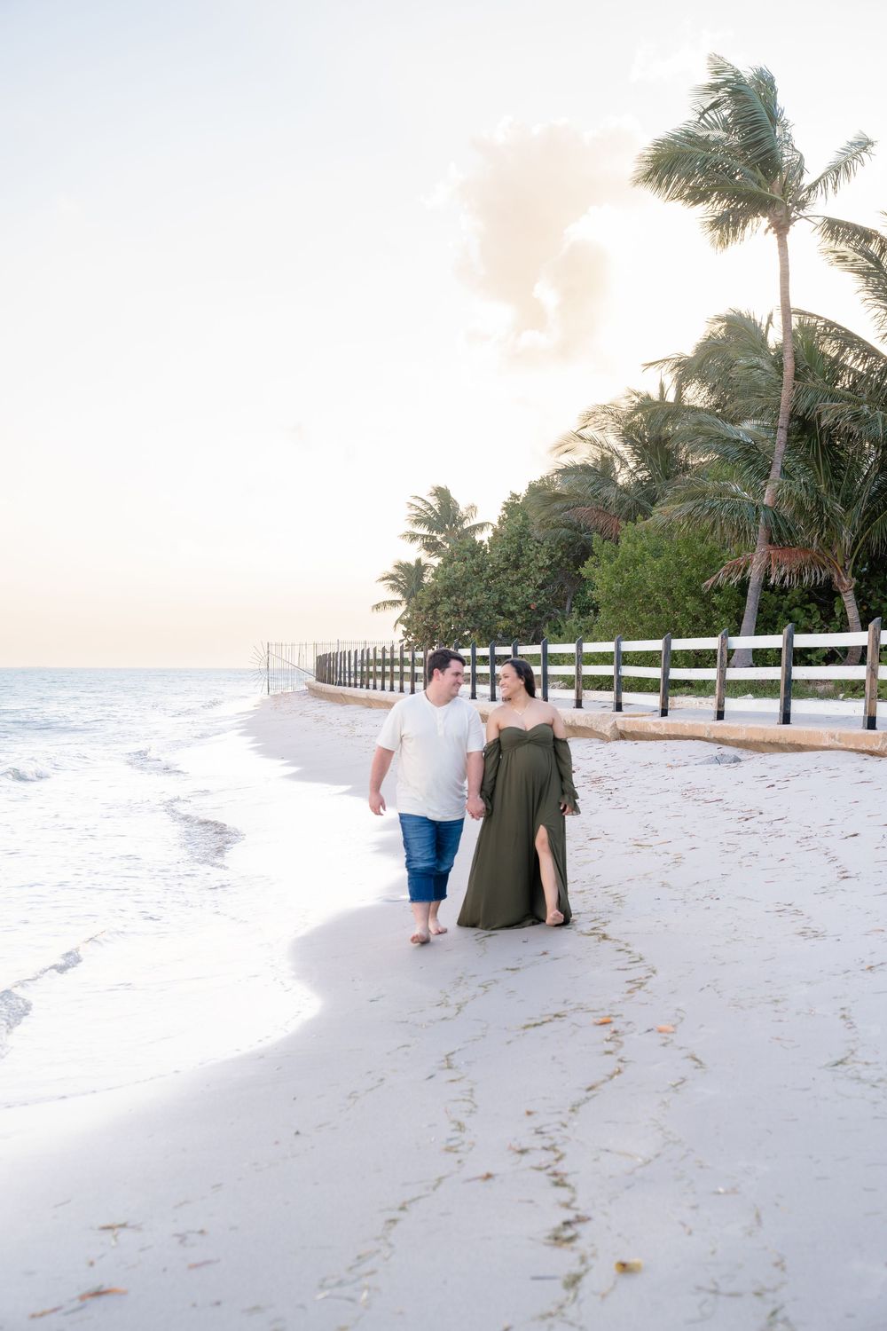 Couple walking hand in hand along a pristine beach boardwalk with palm trees at sunset.