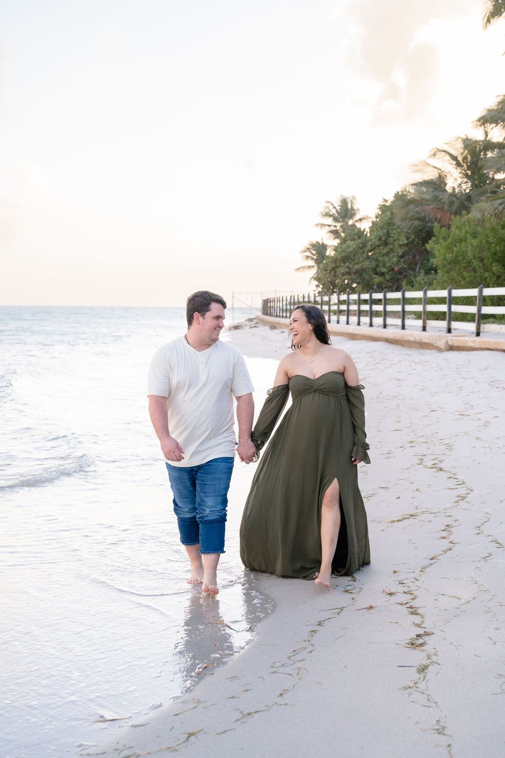 Romantic beach stroll by couple in white shirt and olive dress along shoreline with palm trees.
