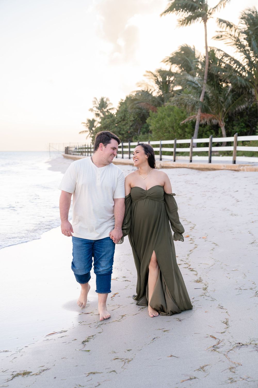 A couple walking barefoot along a white sandy beach with palm trees in the background during sunset.