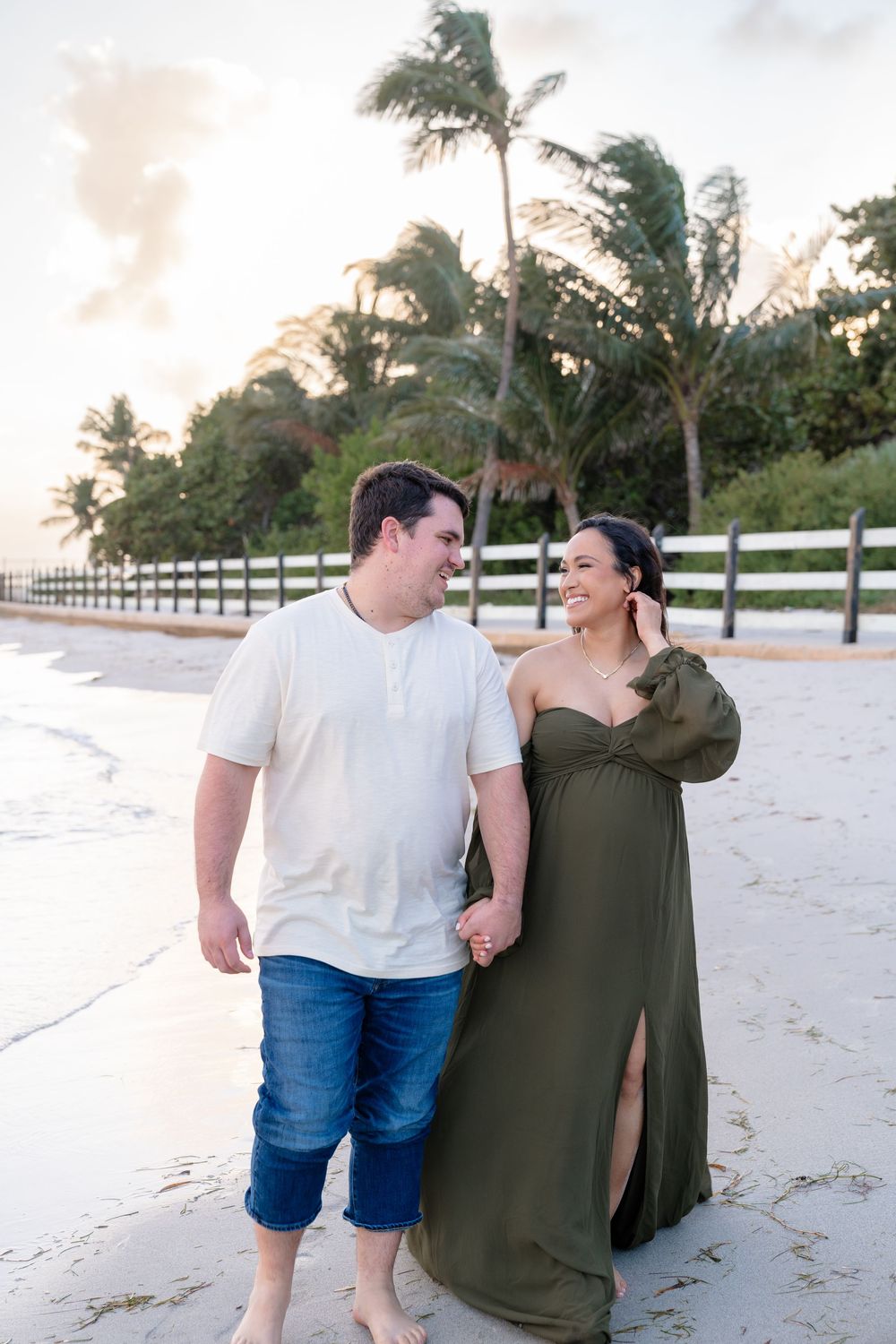 A romantic beach photoshoot with a couple embracing on sandy shores with palm trees at sunset.
