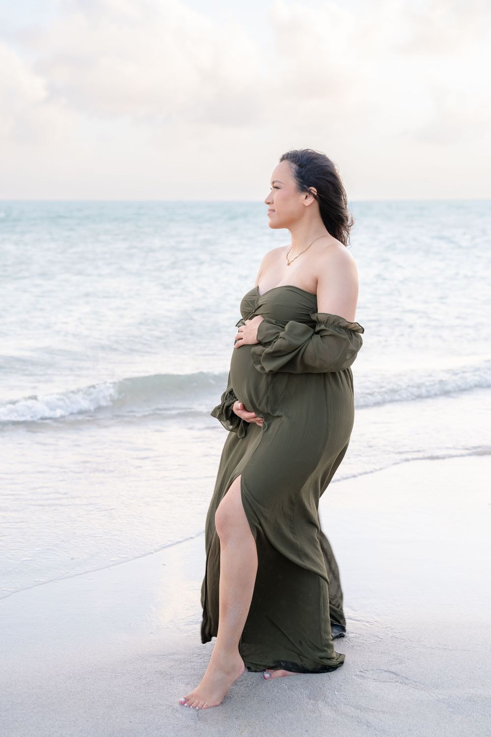 A woman in an olive green off-shoulder dress poses elegantly on a peaceful beach at sunset.