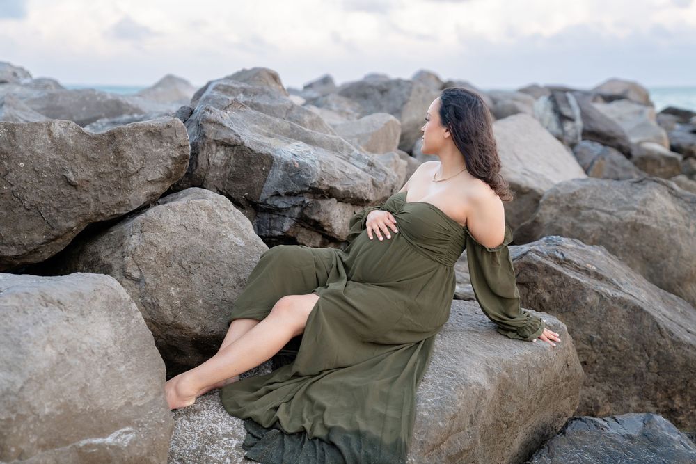 A woman in a flowing olive dress reclines gracefully against coastal rocks near the ocean.
