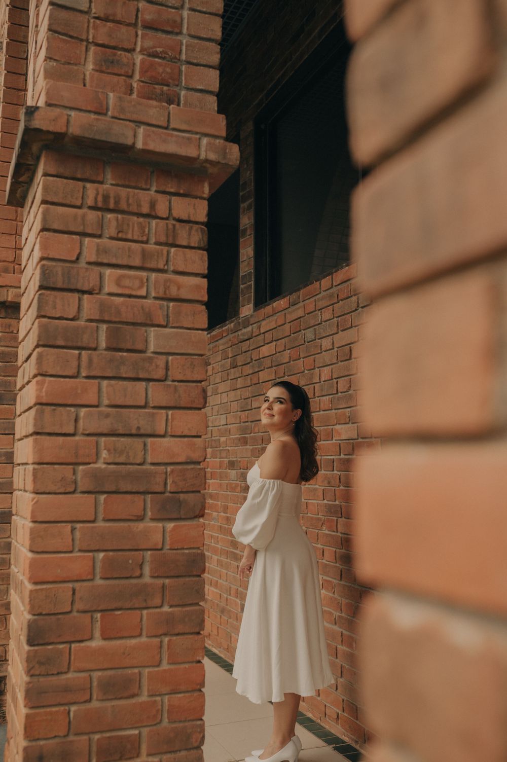 An elegant figure in a white off-shoulder dress poses against a brick wall backdrop.