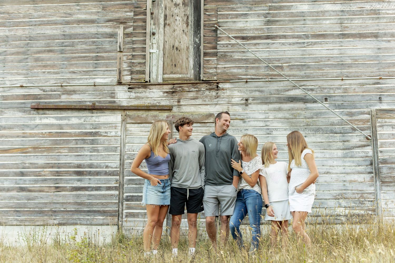 Family poses together against rustic white barn wall in casual summer outfits during outdoor photo session.