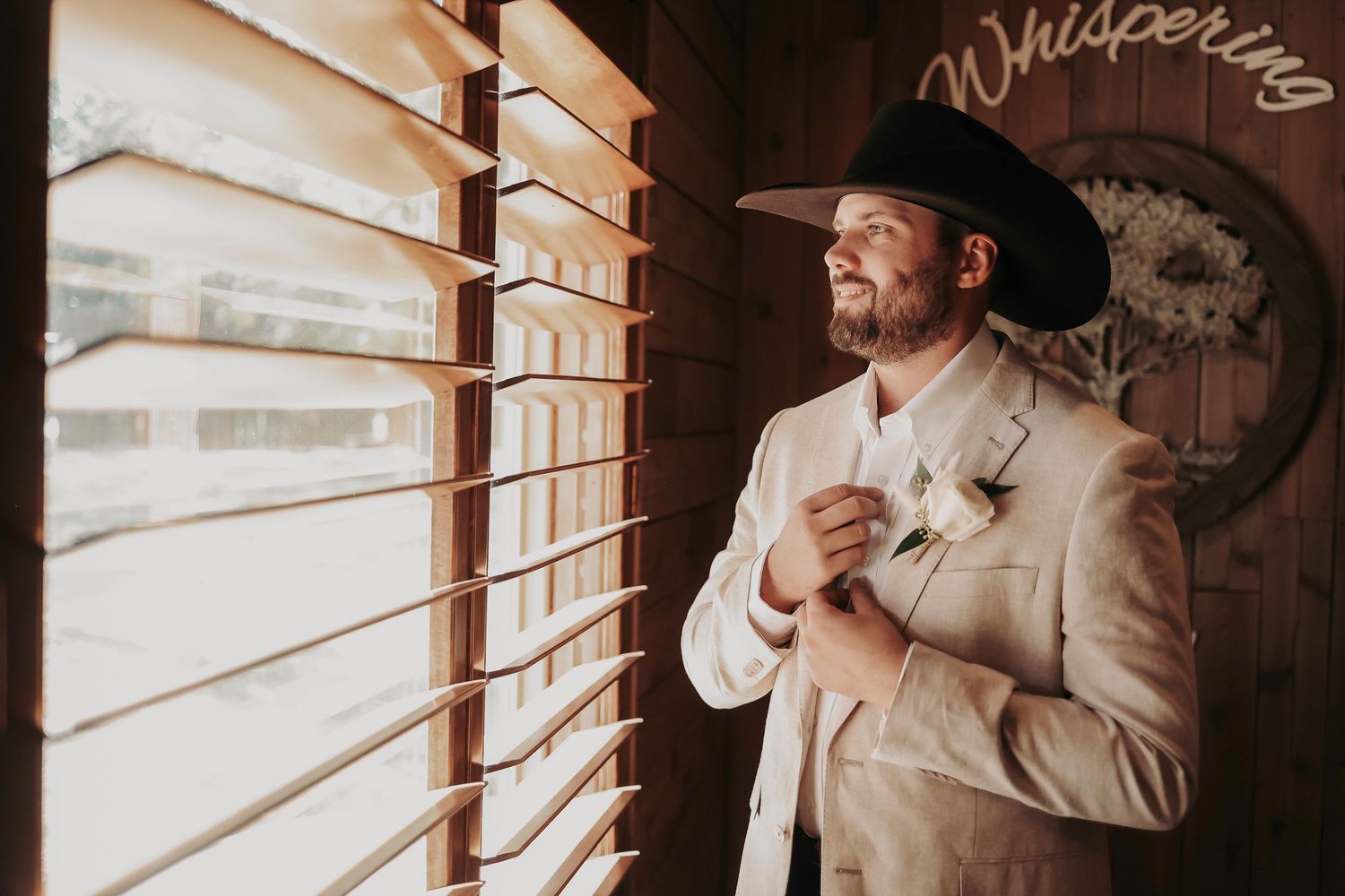 Groom adjusting blazer by rustic barn window at Whispering Oaks