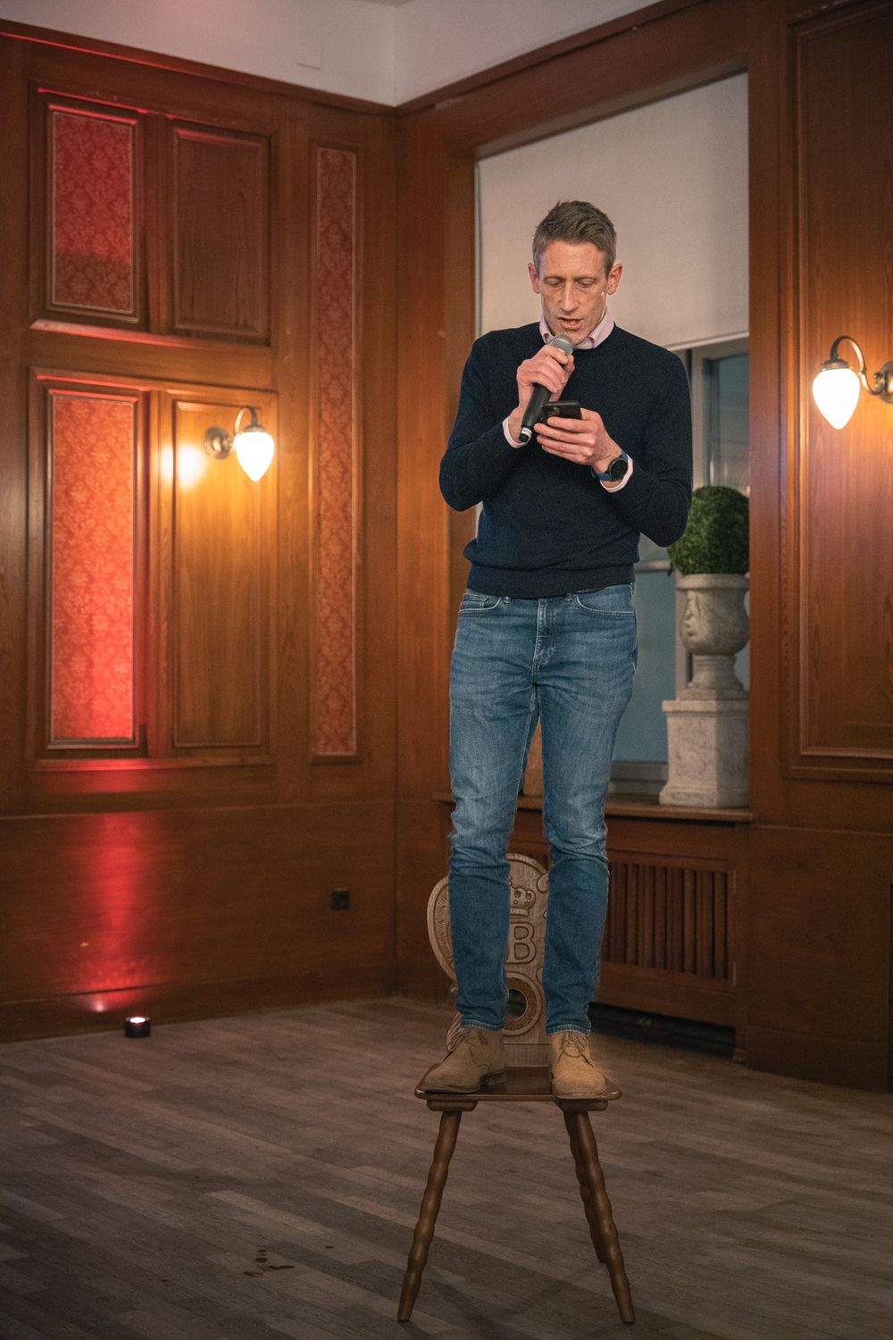 Man in black sweater and jeans stands on wooden stool holding microphone in elegant wood-paneled room.