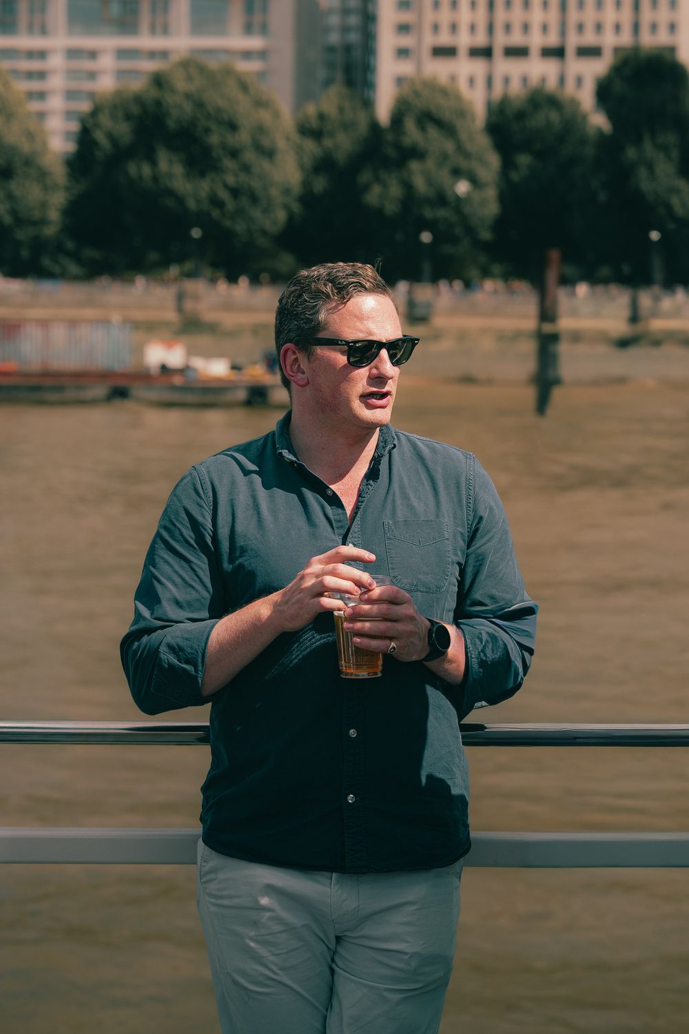 Man in sunglasses holding a beer by a river railing on a sunny day.
