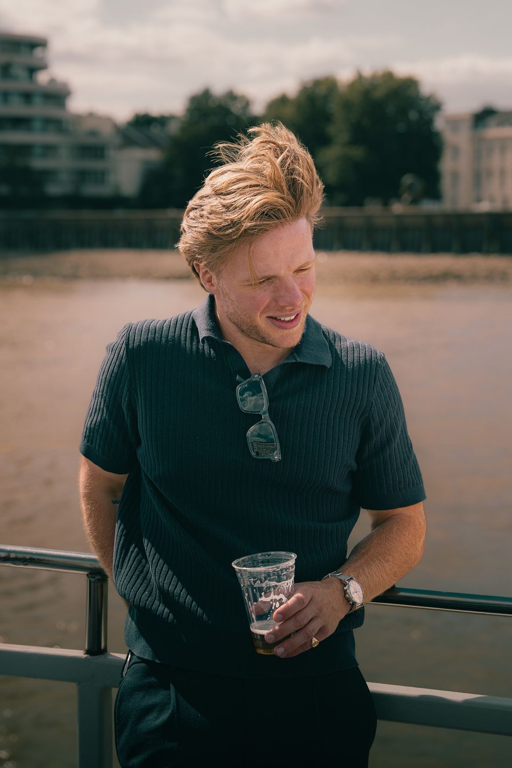Young man with blond hair holding a drink, leaning on a railing by a river, wearing a navy polo shirt.