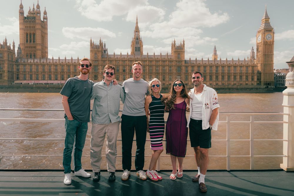 Six friends pose on a riverside deck with the Houses of Parliament and Big Ben in London behind them.