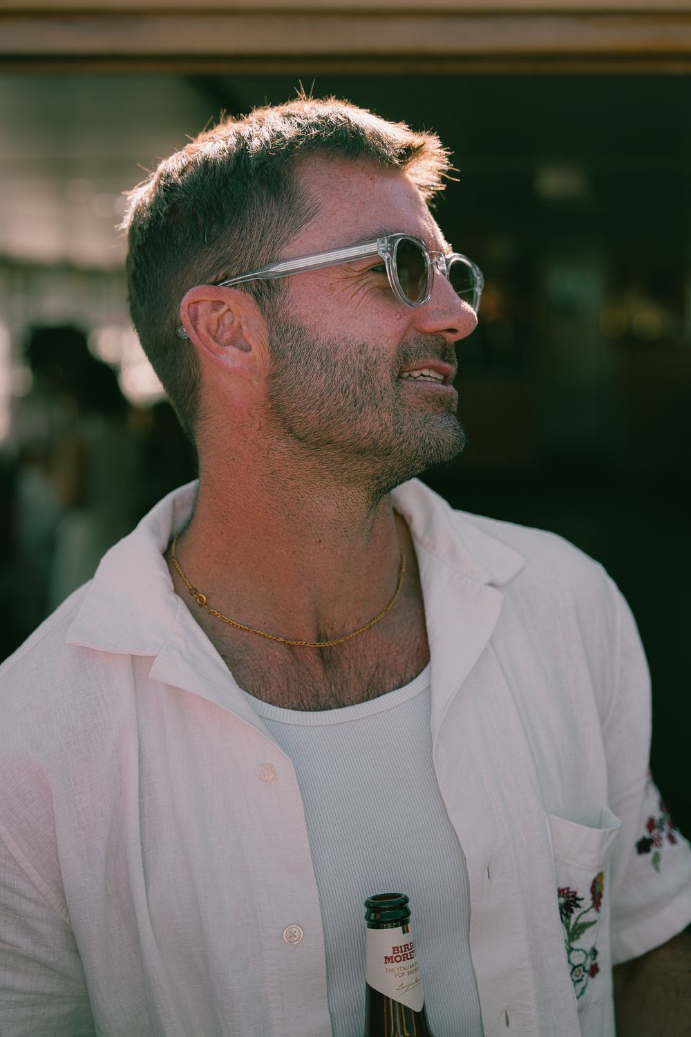 Man in white linen shirt and round sunglasses holding a beer bottle outdoors in warm sunlight.