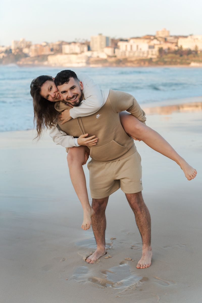 A playful couple enjoys a sunrise moment on Bondi beach with coastal buildings in the background during a Sydney Photoshoot with a couple
