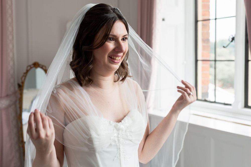A bride in a white wedding dress smiles while adjusting her veil near a window.