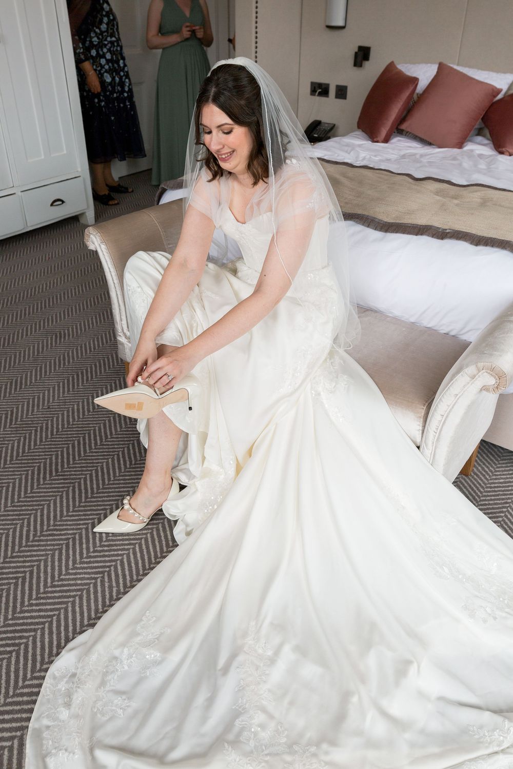 A bride in a flowing white gown sits in a chair while putting on her wedding shoes.