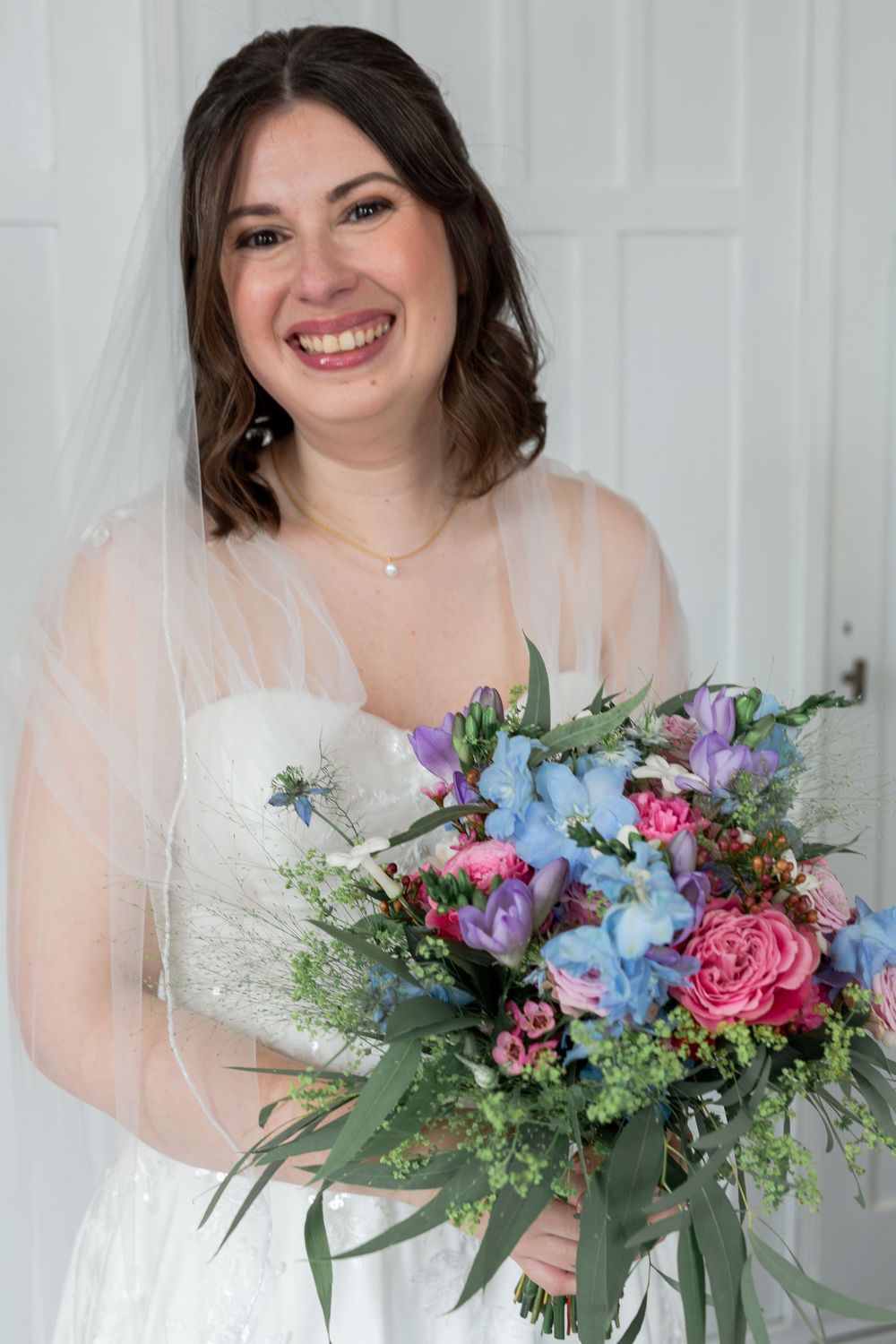 A bride holds a vibrant bouquet featuring blue, pink and purple flowers with greenery.