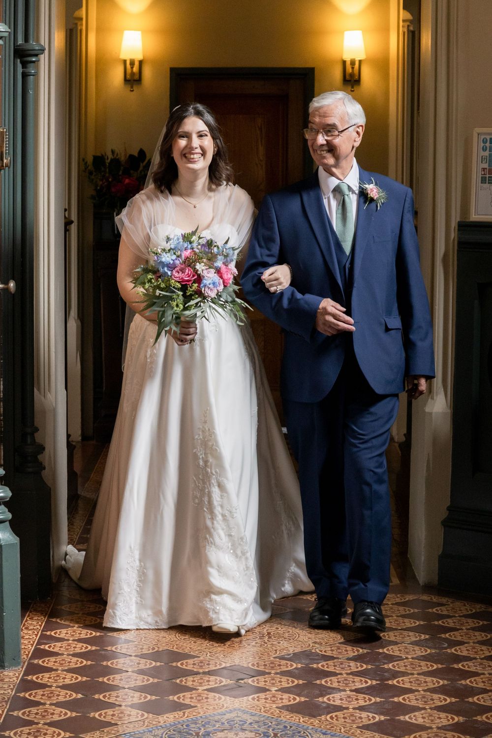 A bride in white dress walks arm in arm with a gentleman in navy suit down an elegant hallway.