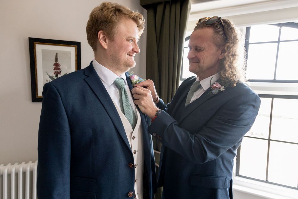 A person adjusts another's tie and boutonniere while getting ready in formal attire before a wedding ceremony.