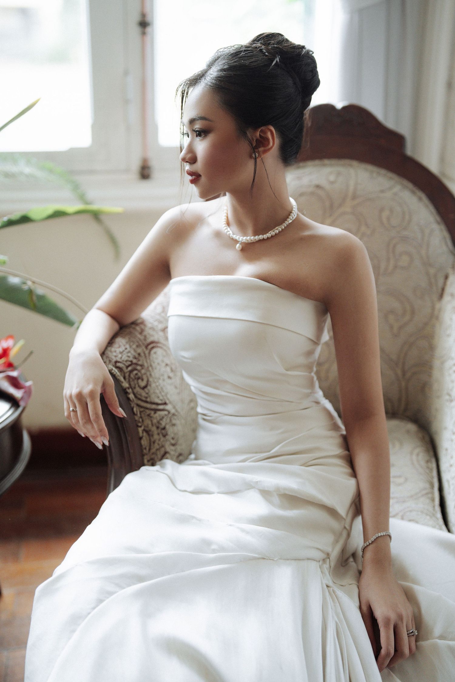 Elegant bride in strapless white gown and pearl necklace sits gracefully on a vintage armchair.