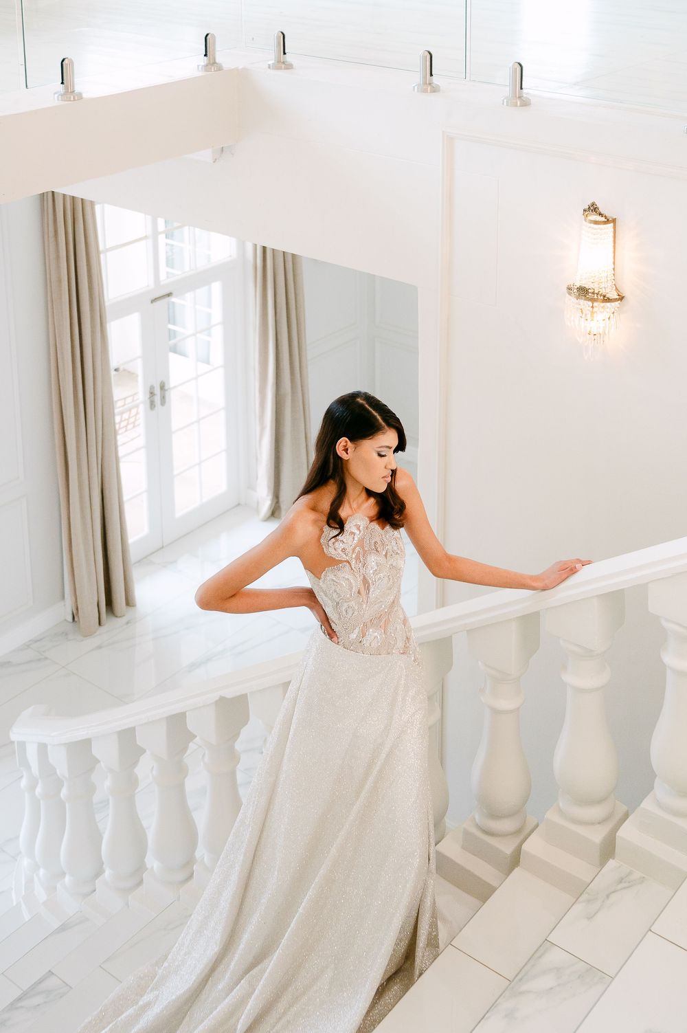 A bride in a flowing white lace gown poses gracefully on an elegant white staircase with natural light streaming in.