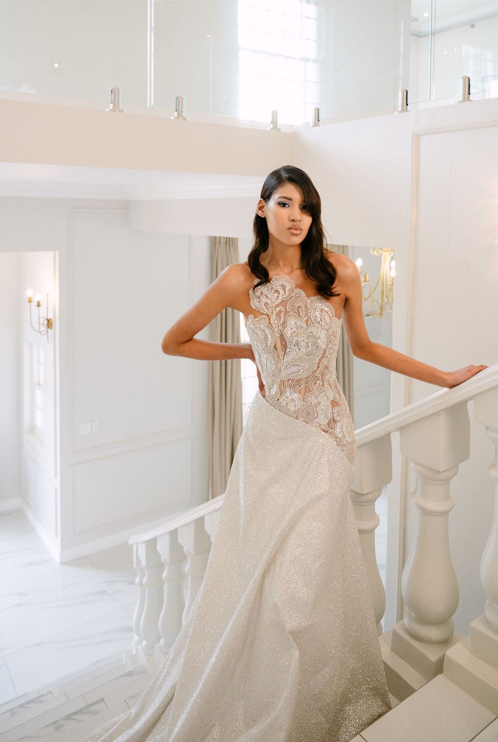 A bride in a romantic lace wedding gown poses elegantly on white stairs in a bright, airy setting.