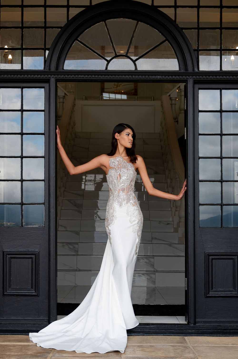 A fitted wedding dress with delicate lace detailing and flowing train is showcased against industrial black windows.