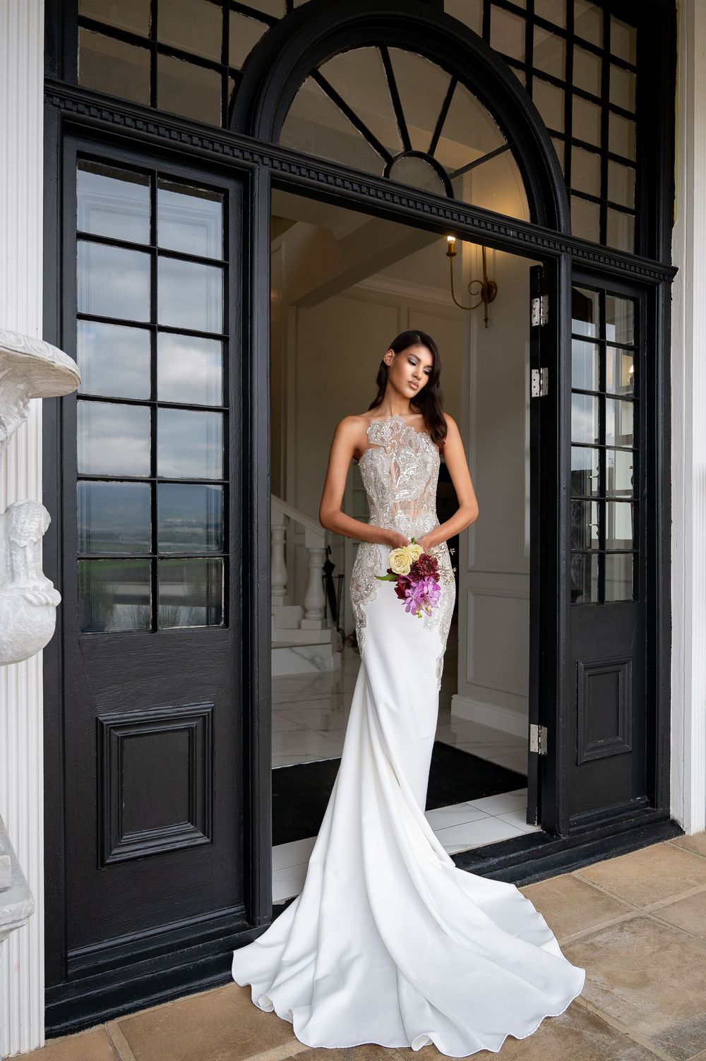 Stunning bridal gown with beaded bodice and flowing train photographed in front of grand entrance with black doors.