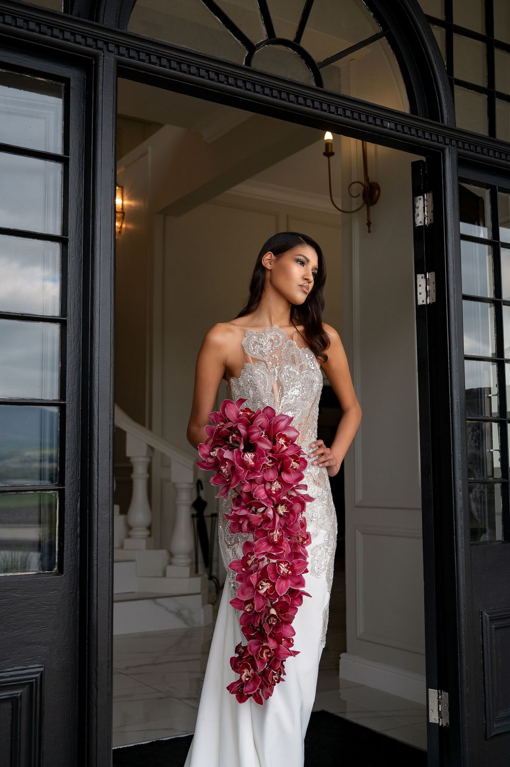 Dramatic bridal portraits in front of black-framed windows holding cascading pink orchid bouquet.