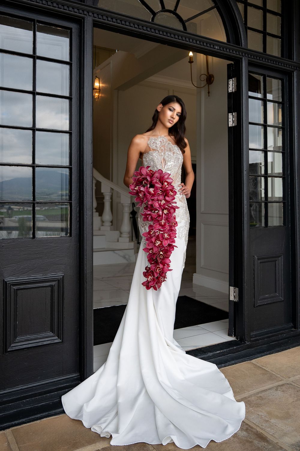 A bride in a flowing white wedding dress poses elegantly with a cascading pink orchid bouquet at a chic black-framed venue.