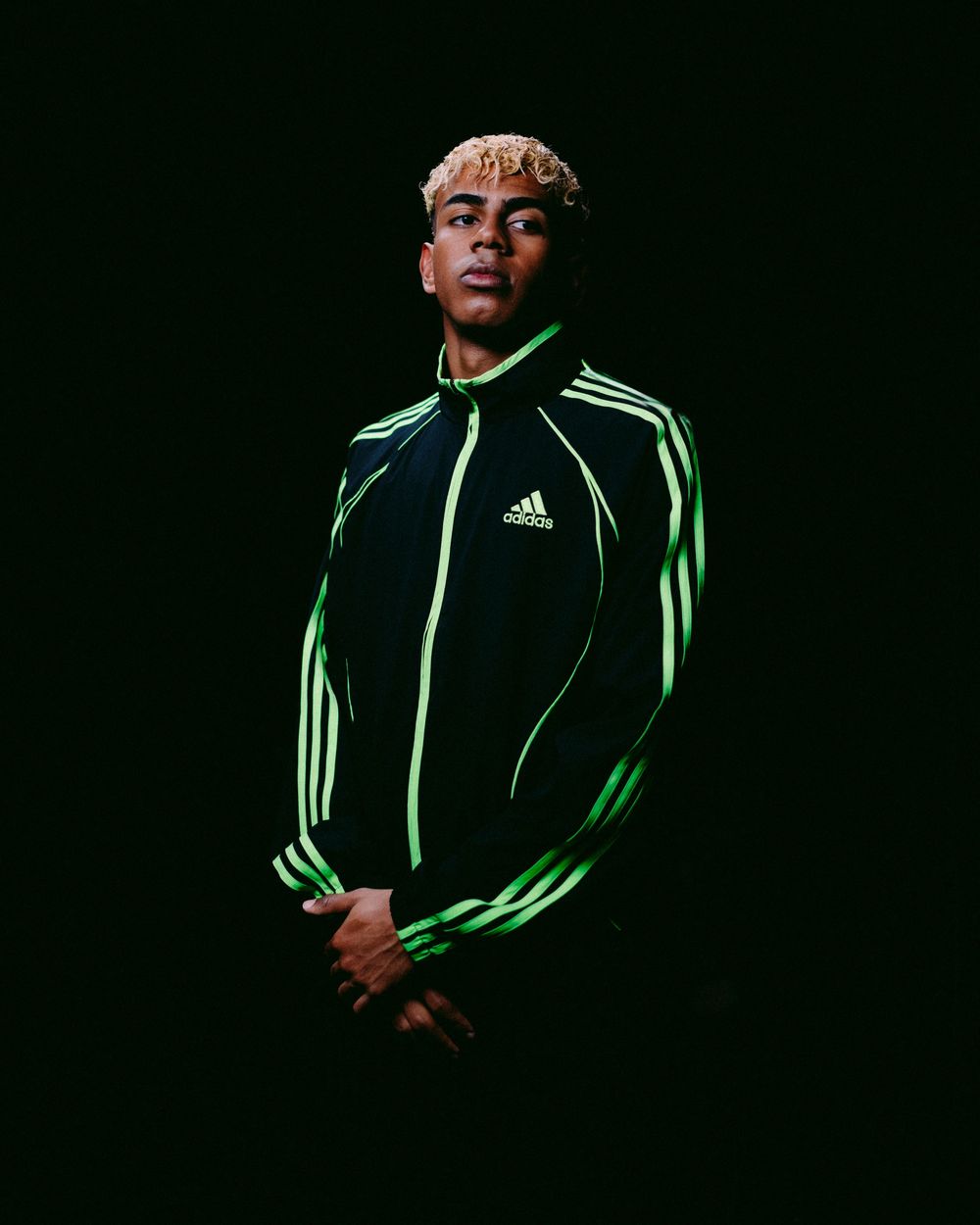 Athletic figure in black and neon green Adidas tracksuit poses dramatically against dark background.