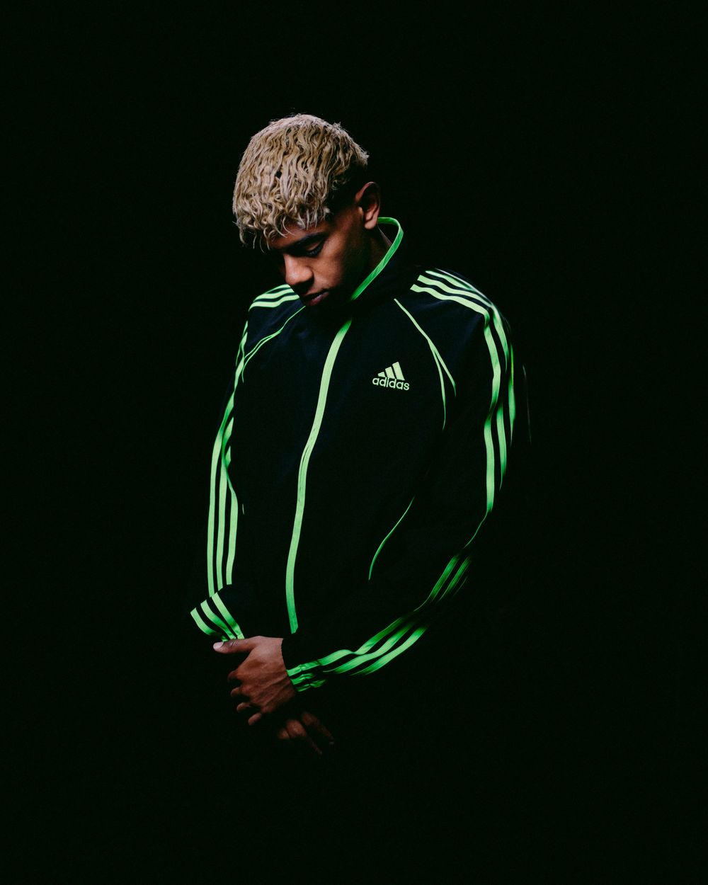 Person in black Adidas track jacket with green stripes poses against dark background in dramatic lighting.