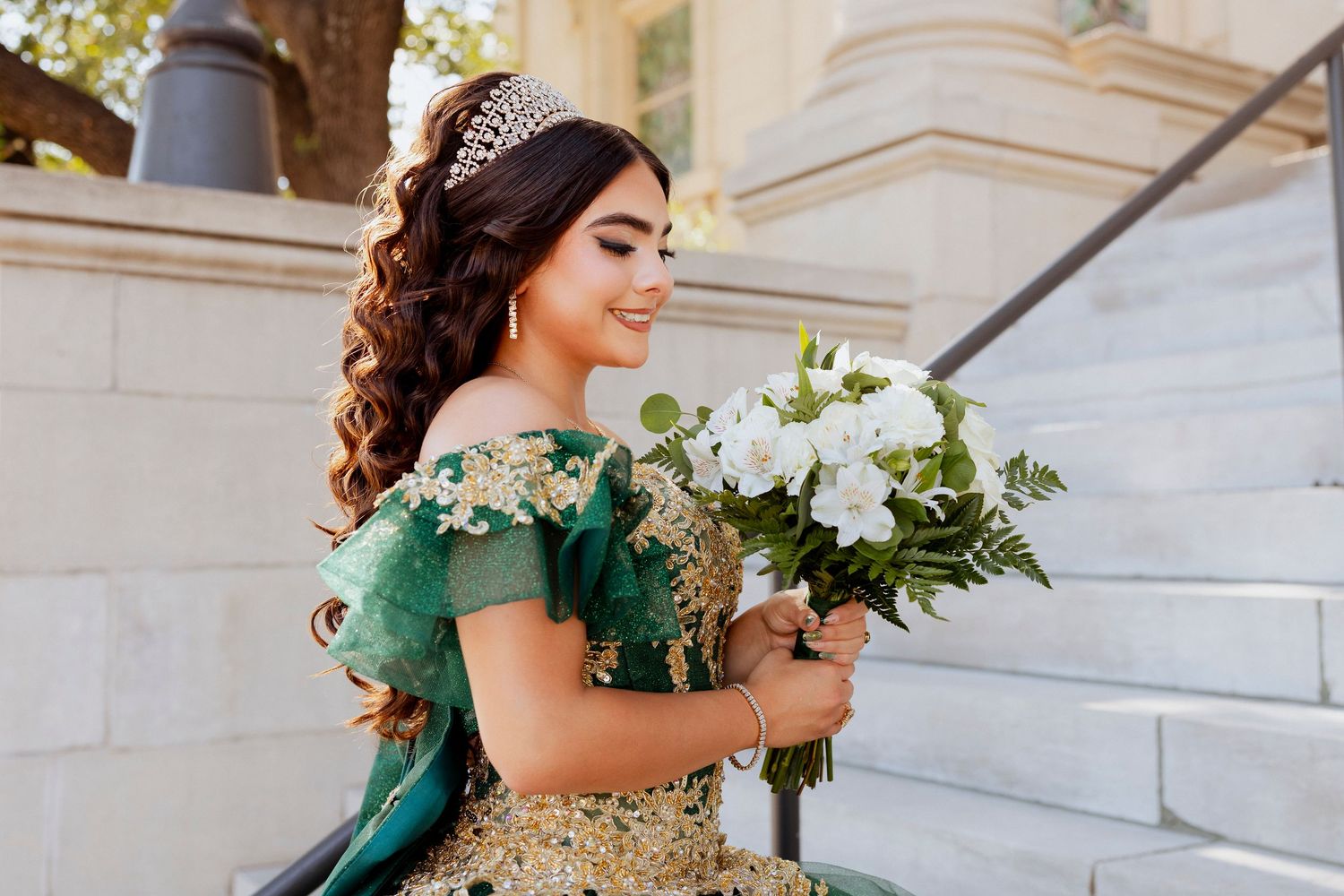 A person in an emerald green gown holds a white floral bouquet while standing on marble steps.