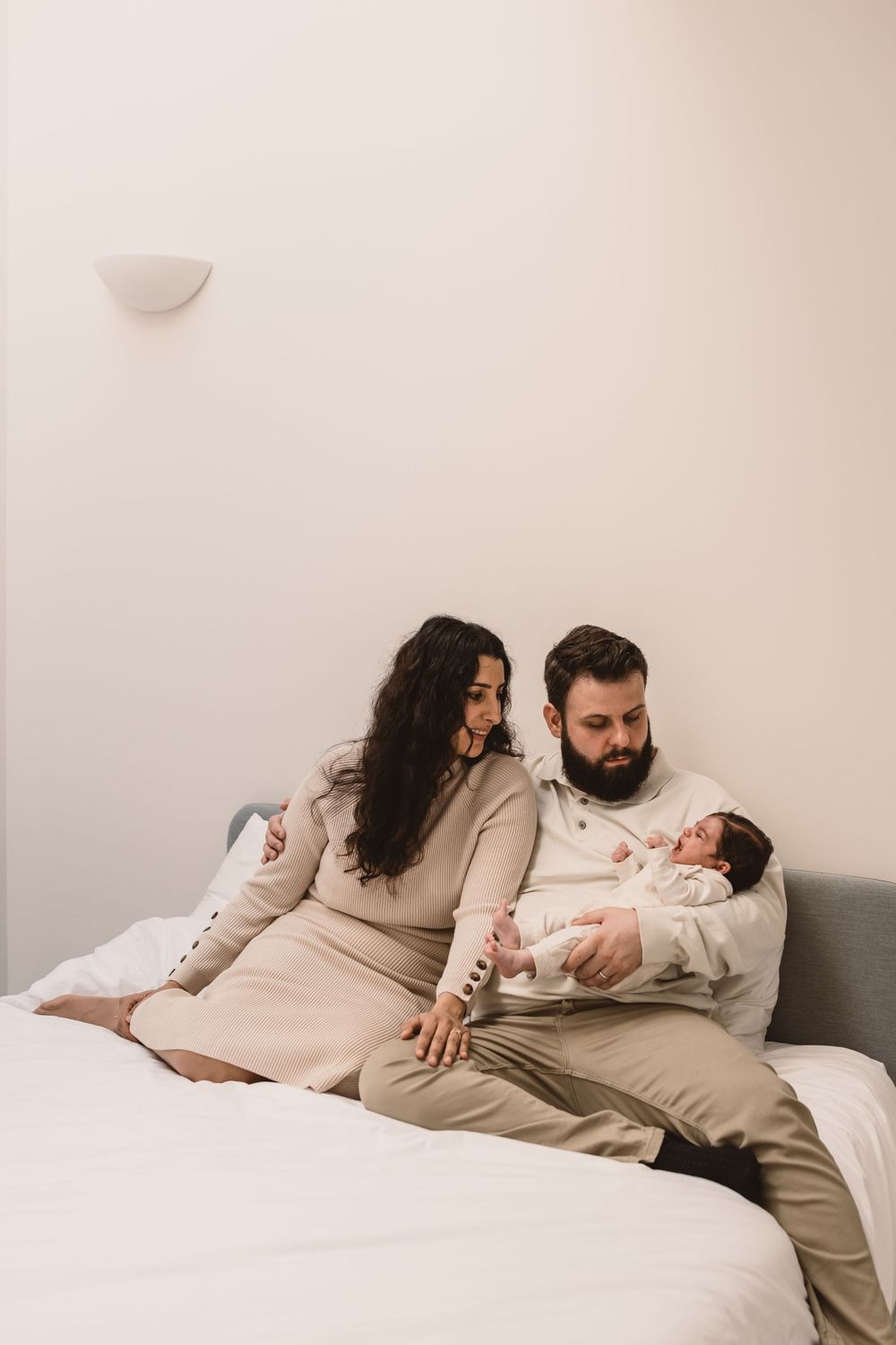 A couple in beige clothing relaxes together on a white bed holding their baby.