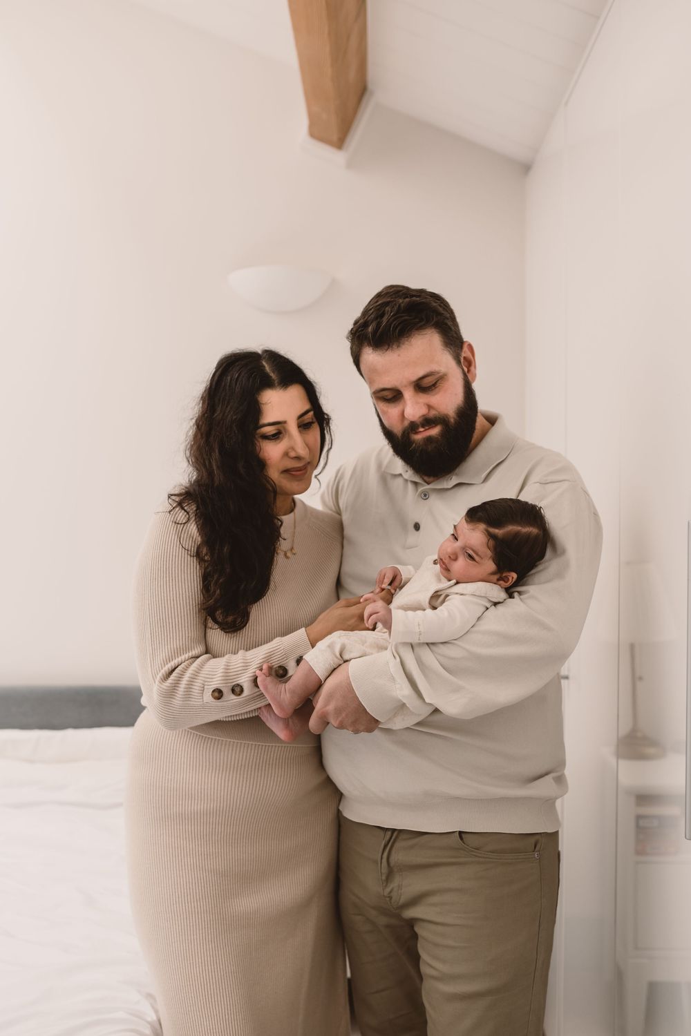 A couple in neutral-toned clothing tenderly hold their newborn baby in a minimalist white bedroom setting.