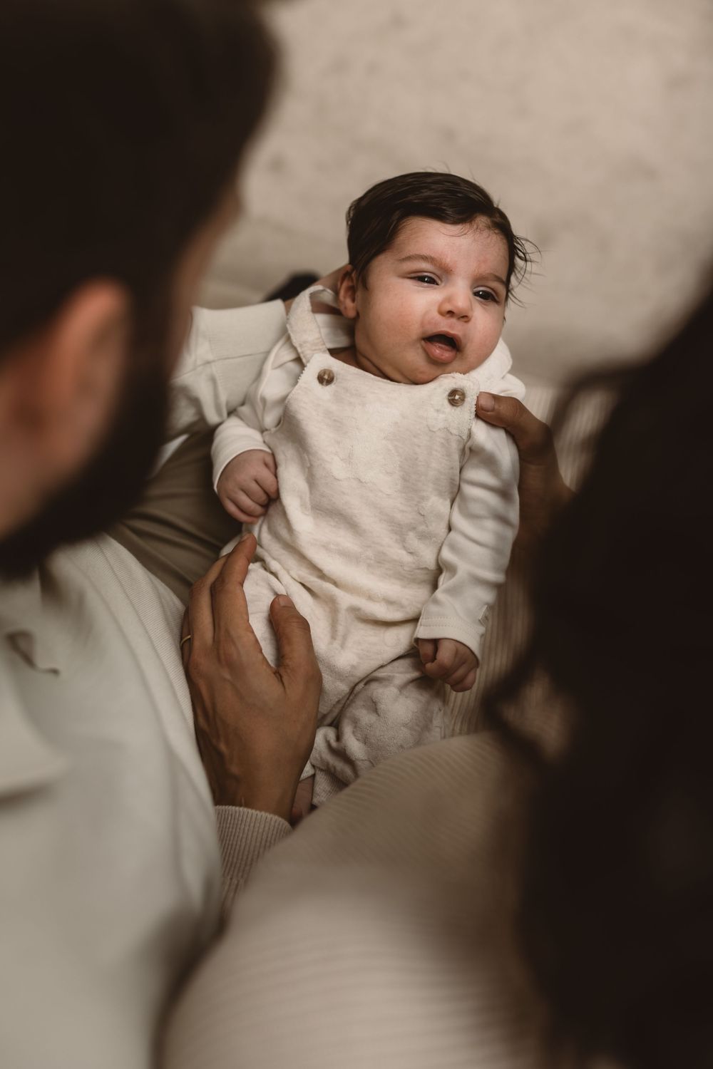 A baby in a white onesie being held and cradled in parents' arms with a blurred background.
