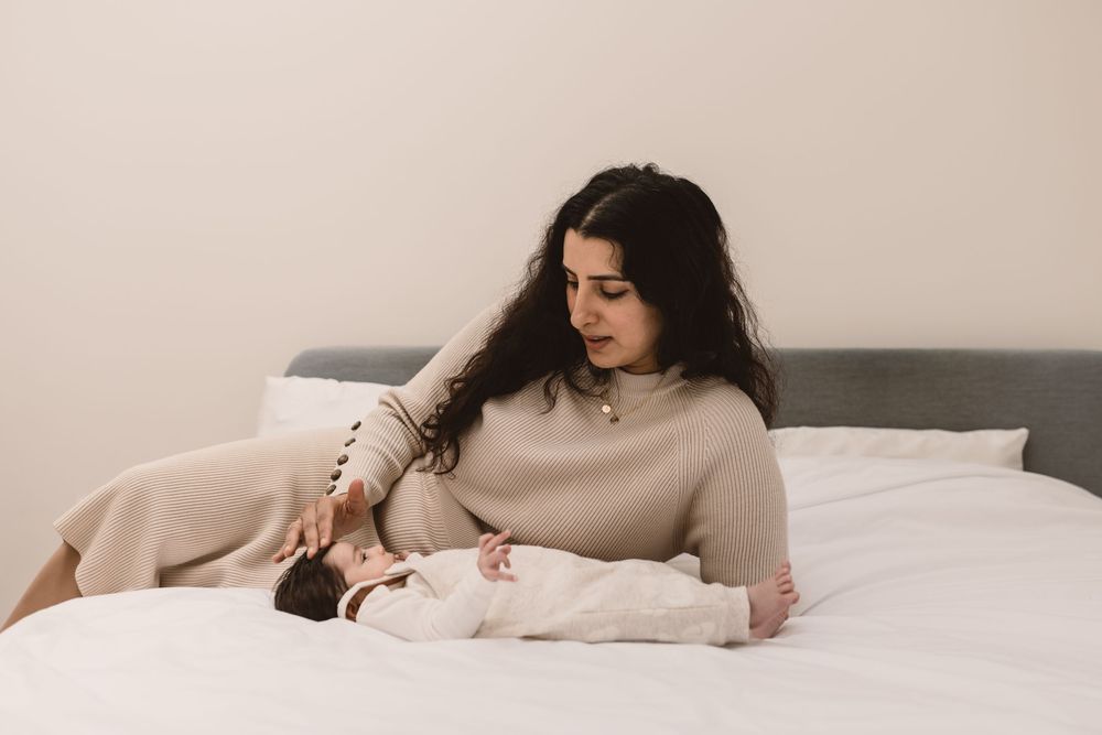 Mother in a beige dress lies on a white bed while looking down at a small baby.
