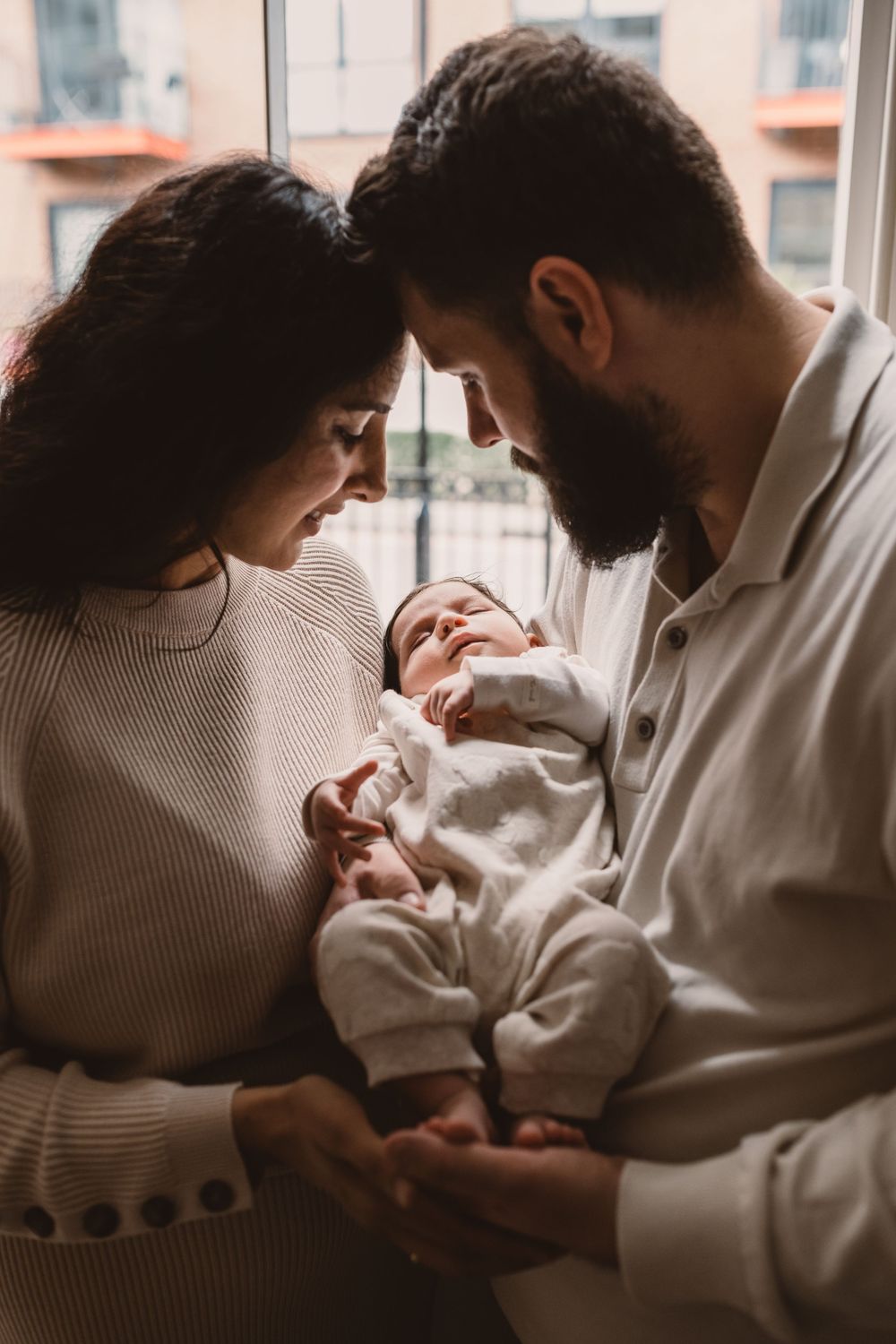 An intimate moment captured between parents and baby by a window in soft natural light.