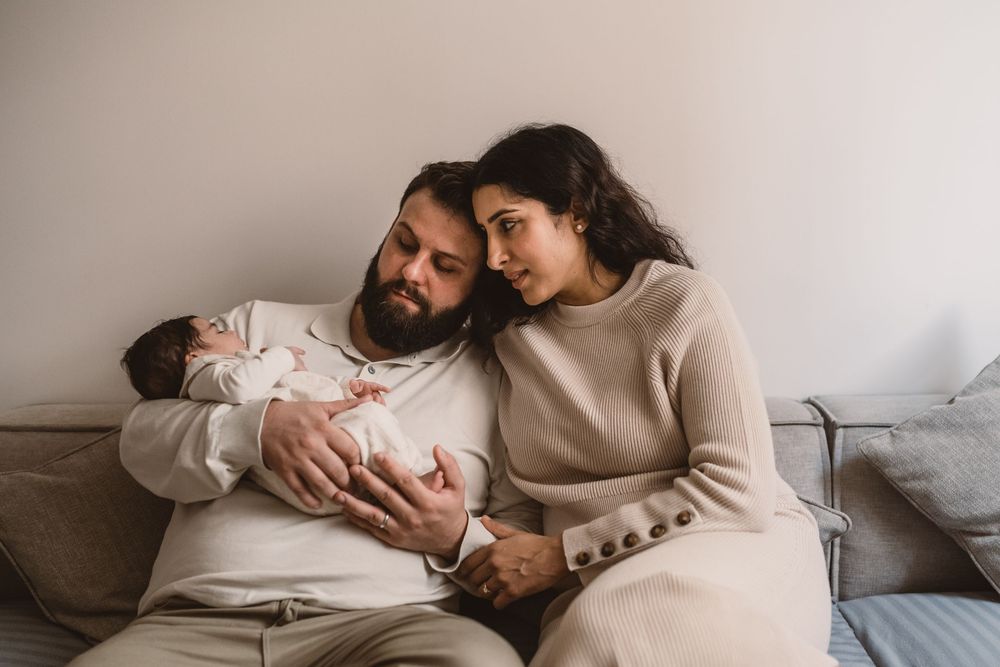 A couple in neutral sweaters sits close together on a couch while holding and gazing at their newborn baby.
