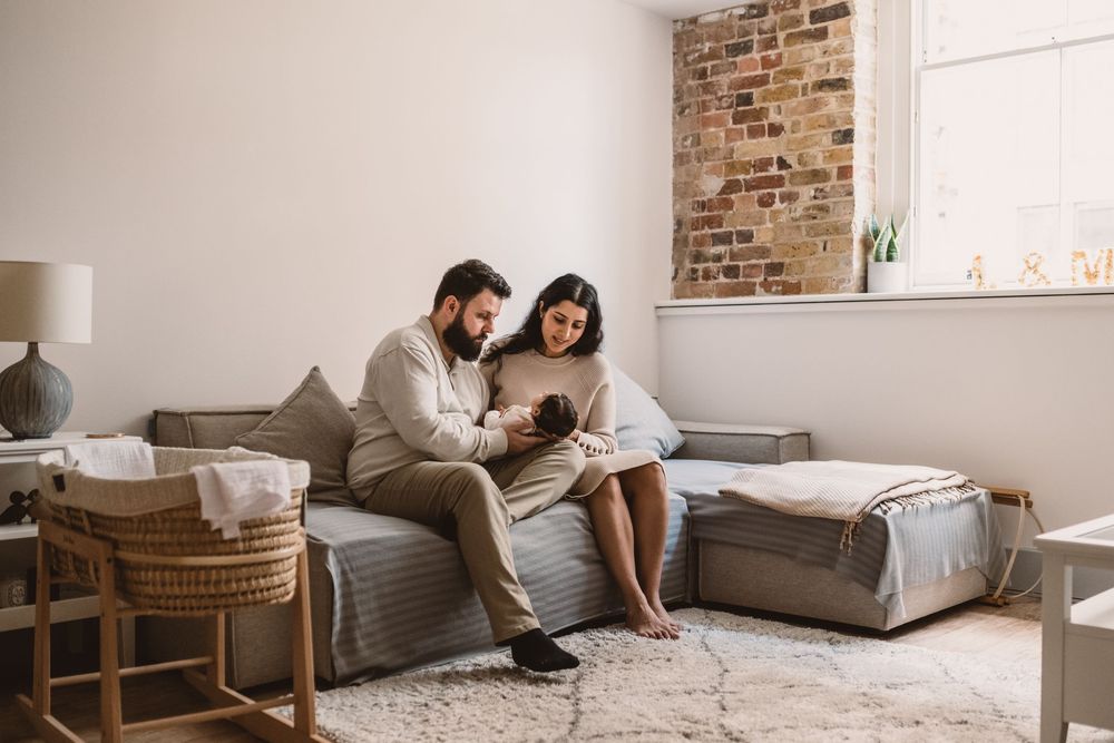 A couple holding their newborn baby relaxes on a gray couch in a modern living room with exposed brick wall.