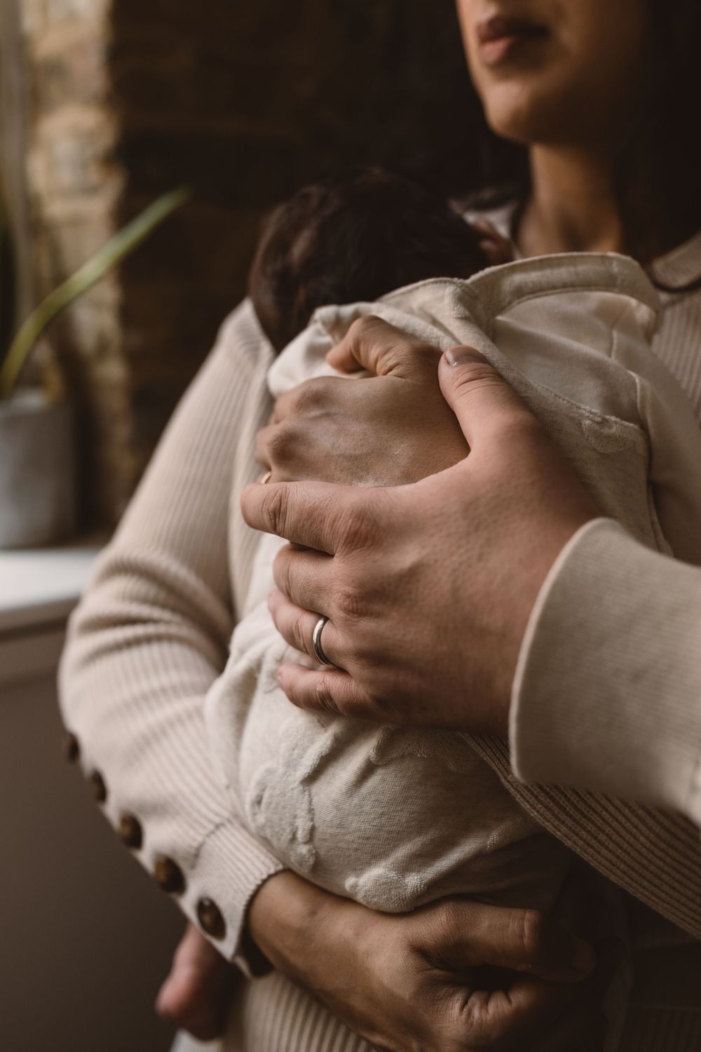 Gentle hands holding and cradling a small infant wrapped in soft white clothing in warm lighting.