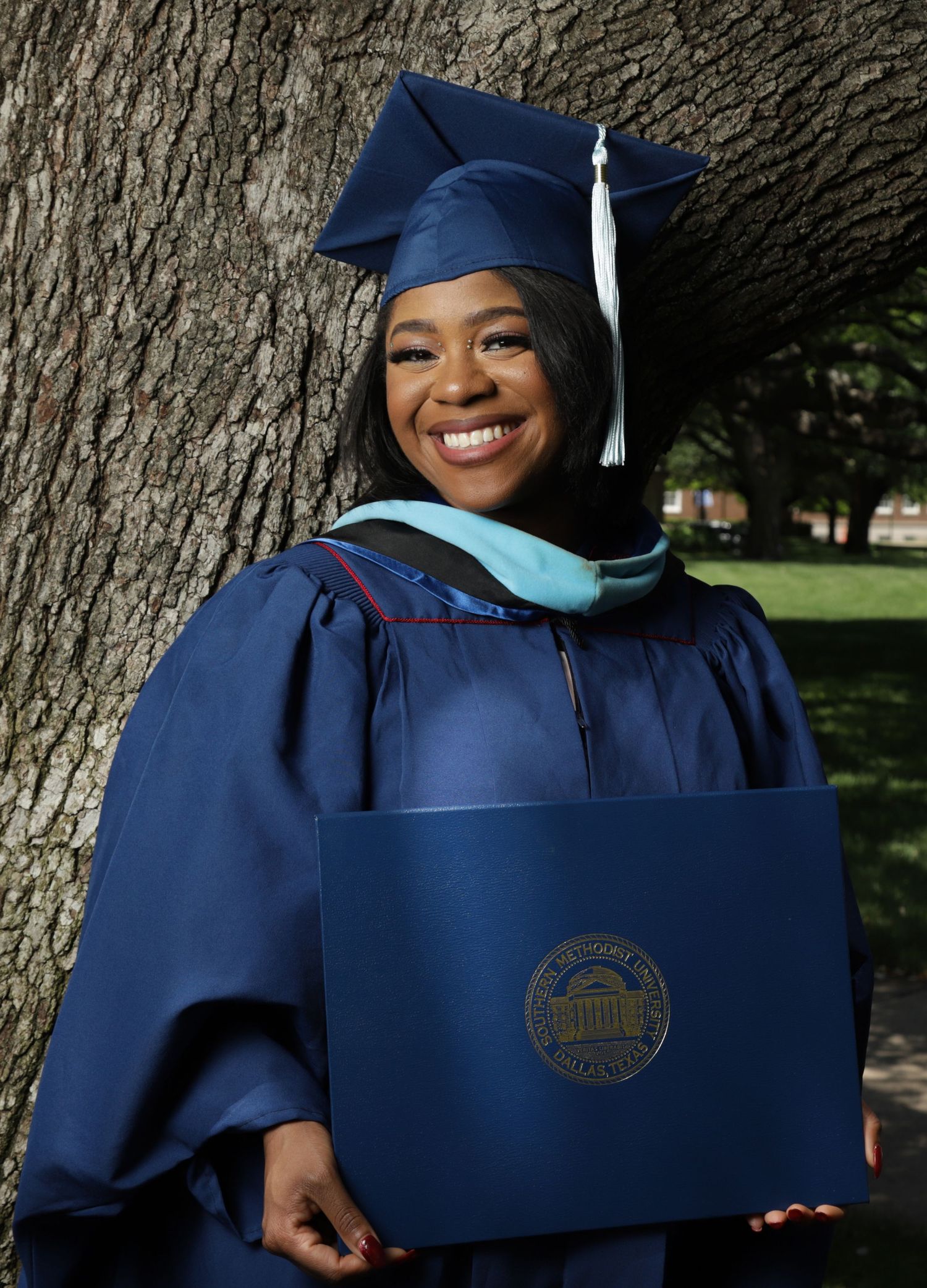 Graduate in navy blue academic regalia smiles and poses with diploma in outdoor setting.