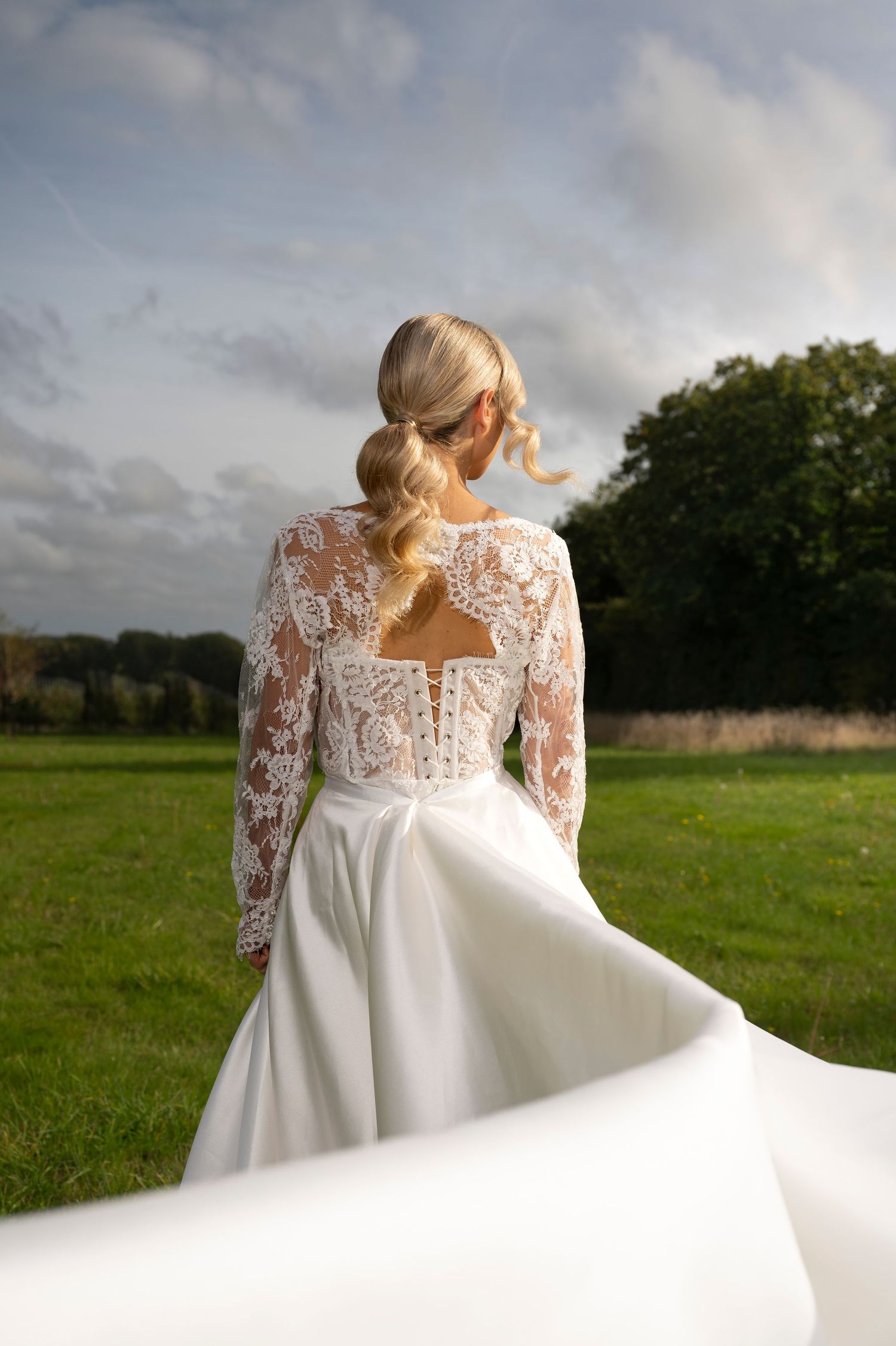 A bride in a flowing white lace gown with long sleeves twirls in a green meadow under cloudy skies.