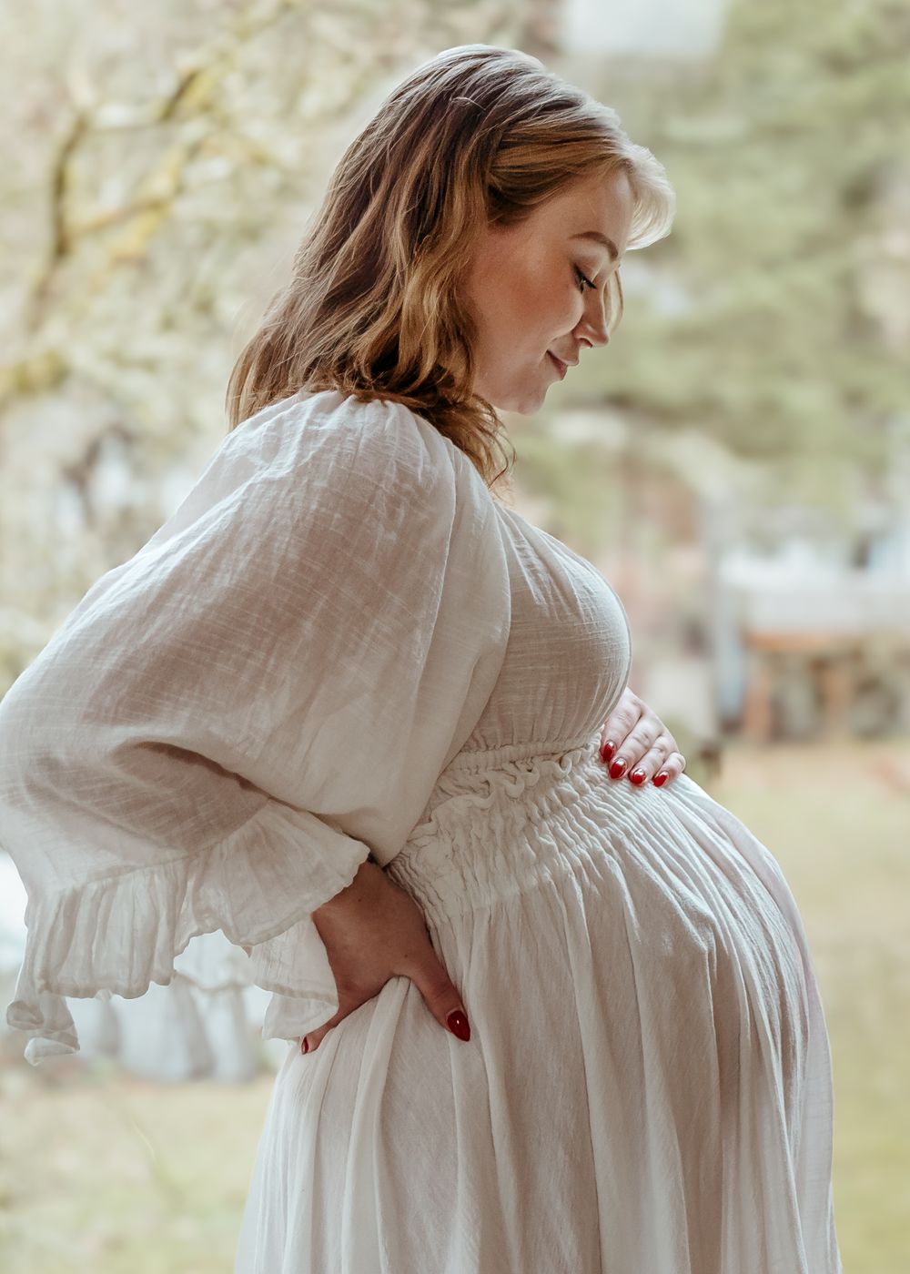 A dreamy maternity portrait shows someone in a white dress touching their baby bump in soft natural light.