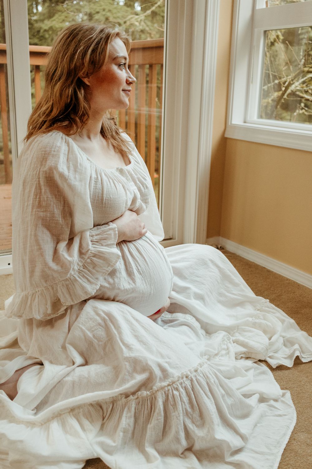 Expectant figure in flowing white cotton dress gazes pensively through window while seated on floor.