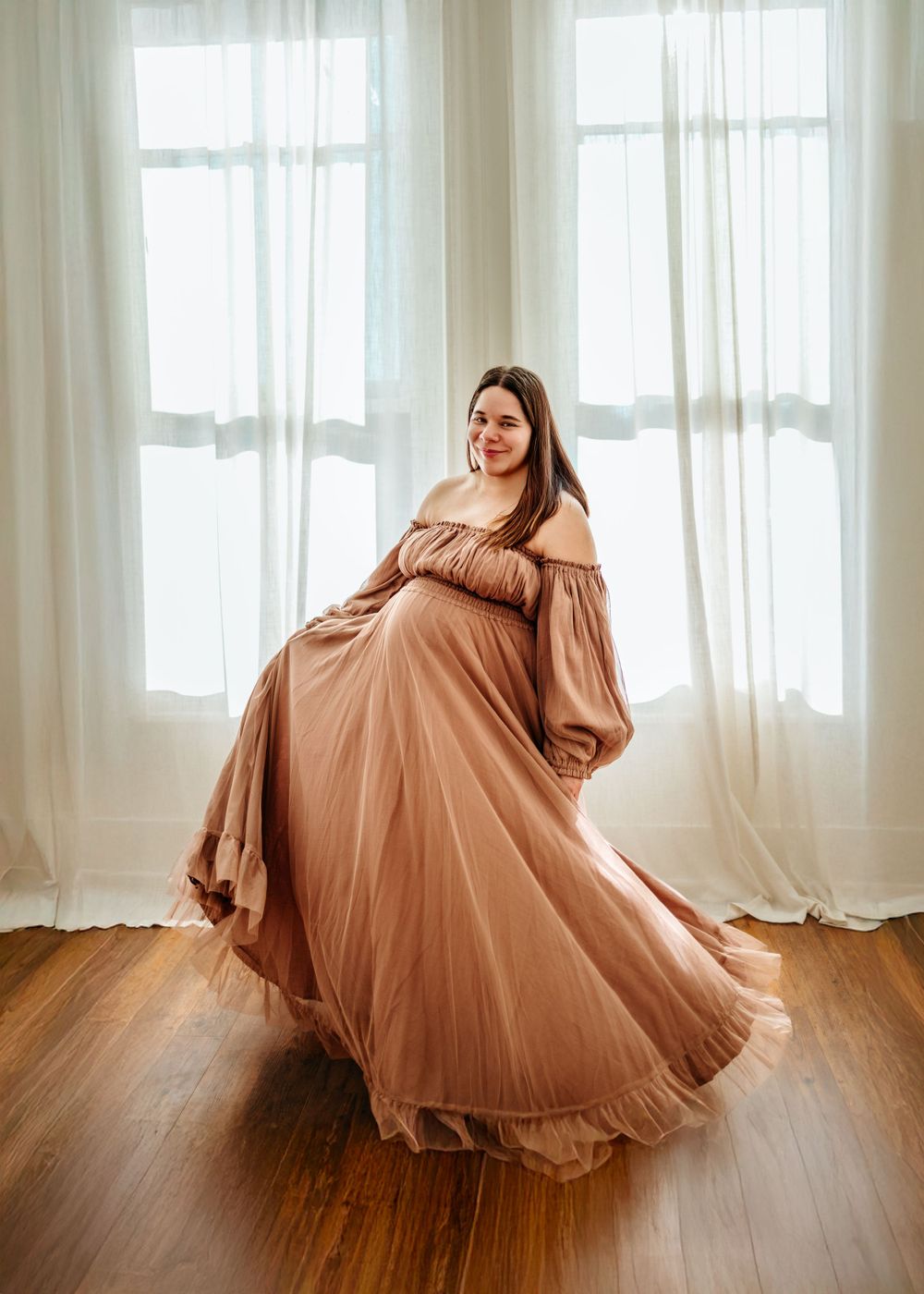 Elegant maternity portrait of woman twirling in flowing mauve gown against bright window.