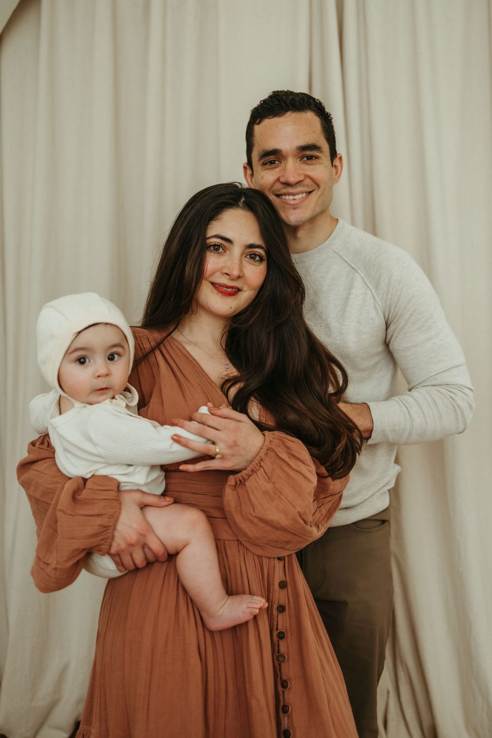 Family portrait with baby in white against cream curtain backdrop.