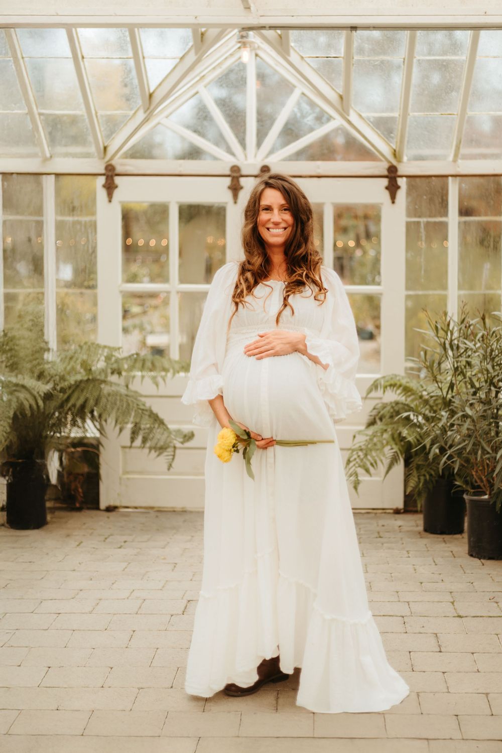 Ethereal maternity portrait in white dress against greenhouse backdrop with ferns and yellow flowers.