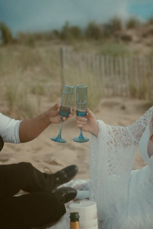 Two people in formal attire hold blue wine glasses while sitting on a sandy beach at sunset.