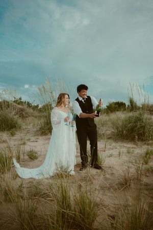 A couple in wedding attire dances together on sand dunes with beach grass and cloudy skies in the background.