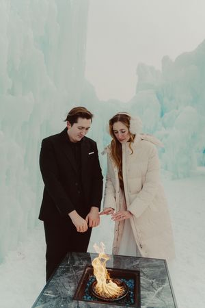 A couple stands beside a fire pit in a snowy winter setting with icy blue-tinted surroundings.