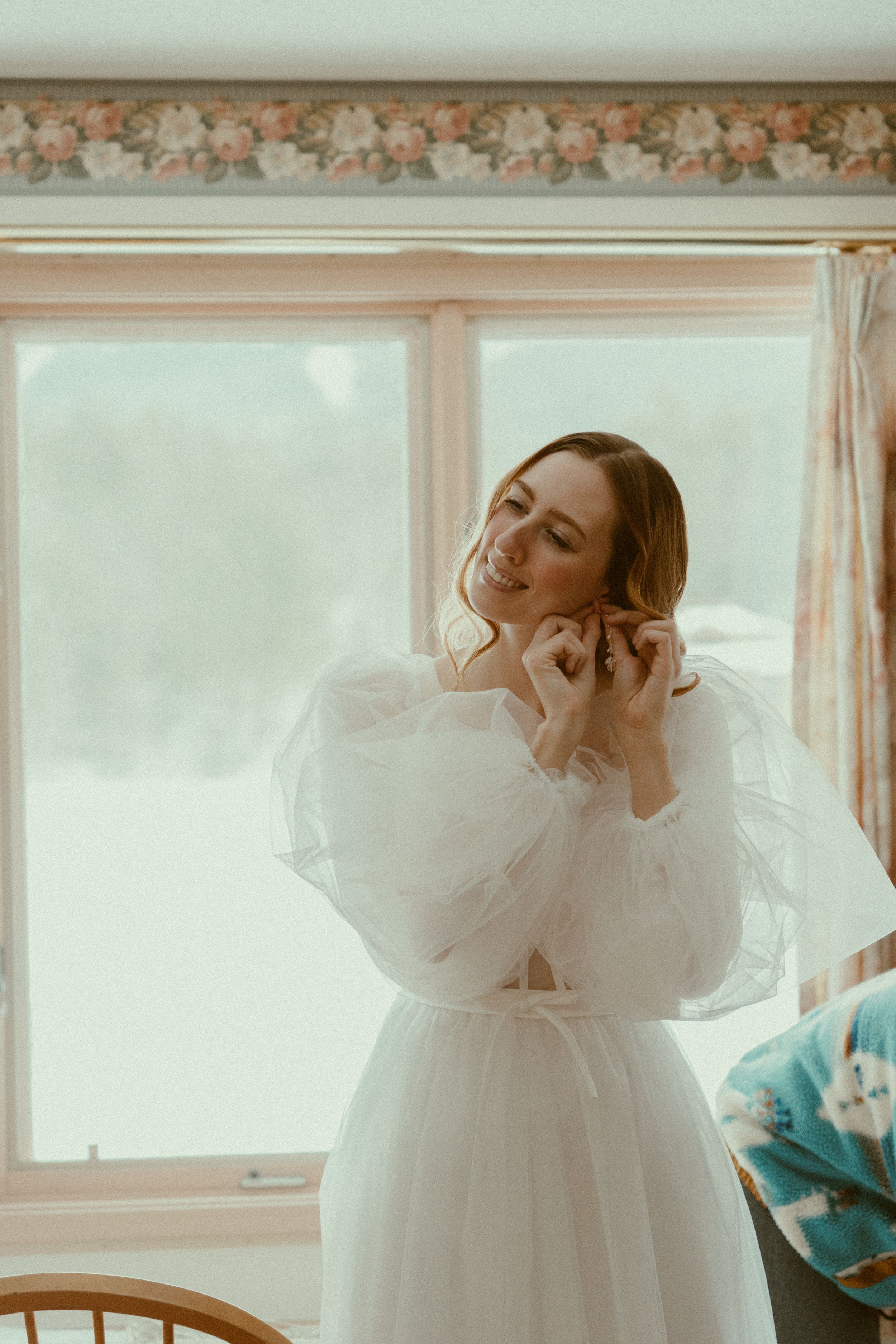 A dreamy portrait of someone in a romantic white tulle dress with billowing sleeves standing by a window in soft light.